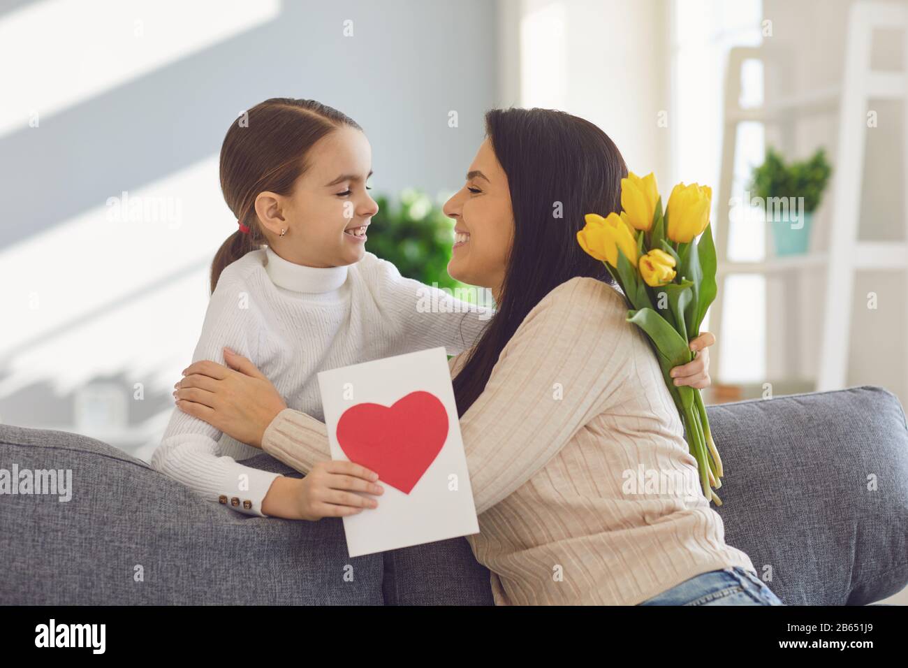 Tag der glücklichen Mütter. Tochter gratuliert Umarmungen, seine Mutter hält einen Blumenstrauß im Zimmer. Stockfoto