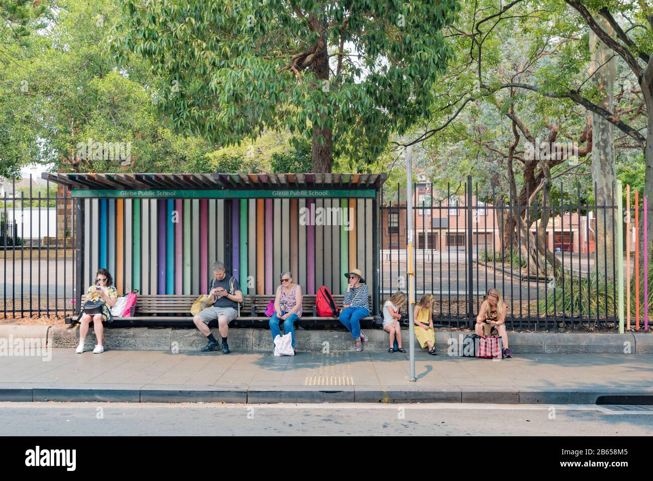 Menschen jeden Alters sitzen und warten an der Bushaltestelle Glebe Public School an einem warmen Sommermorgen auf ihren Bus in Sydney, NSW, Australien Stockfoto