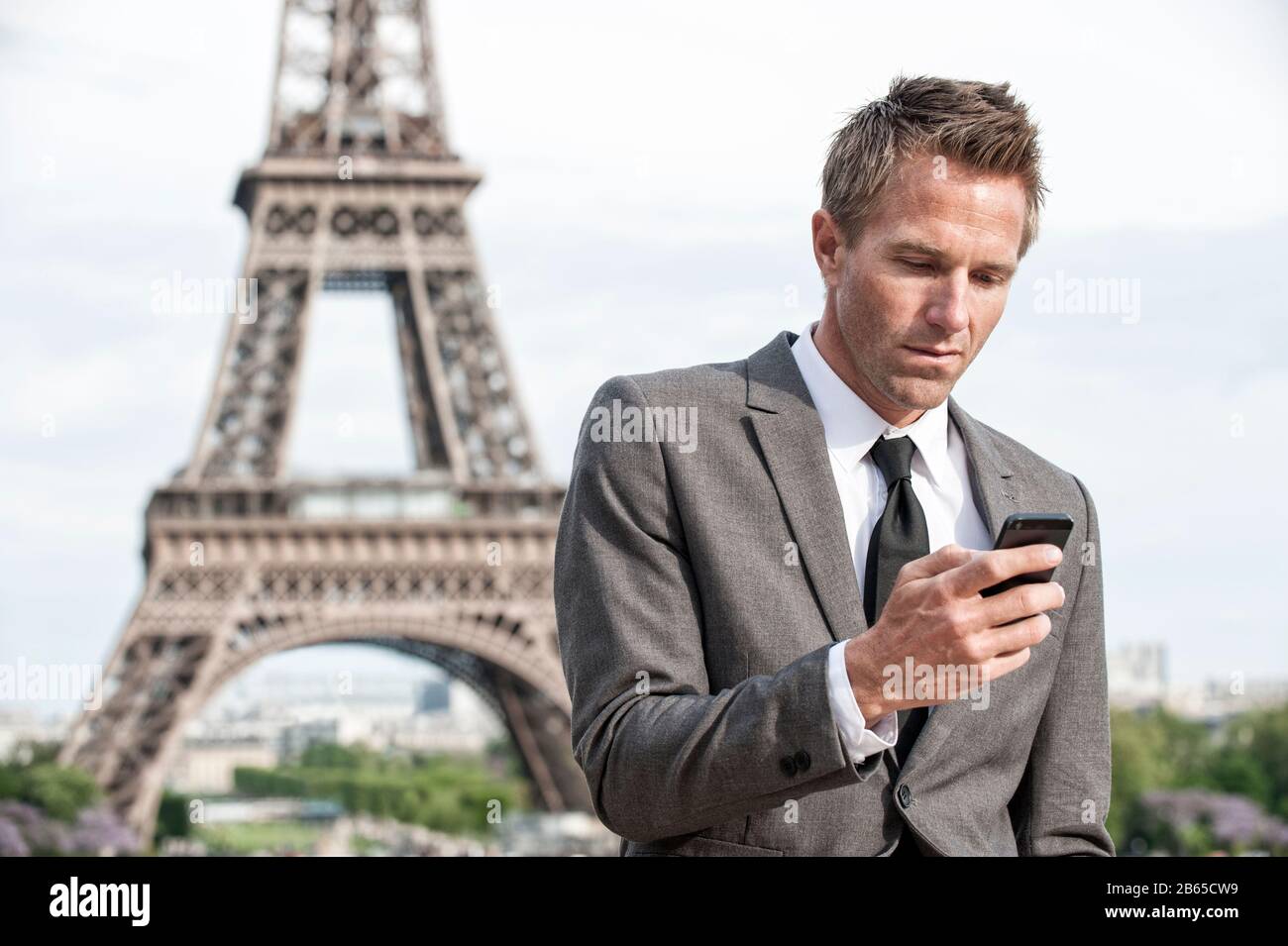 Ein ernsthafter europäischer Geschäftsmann, der im Freien mit seinem Handy vor dem Eiffelturm in Paris steht Stockfoto