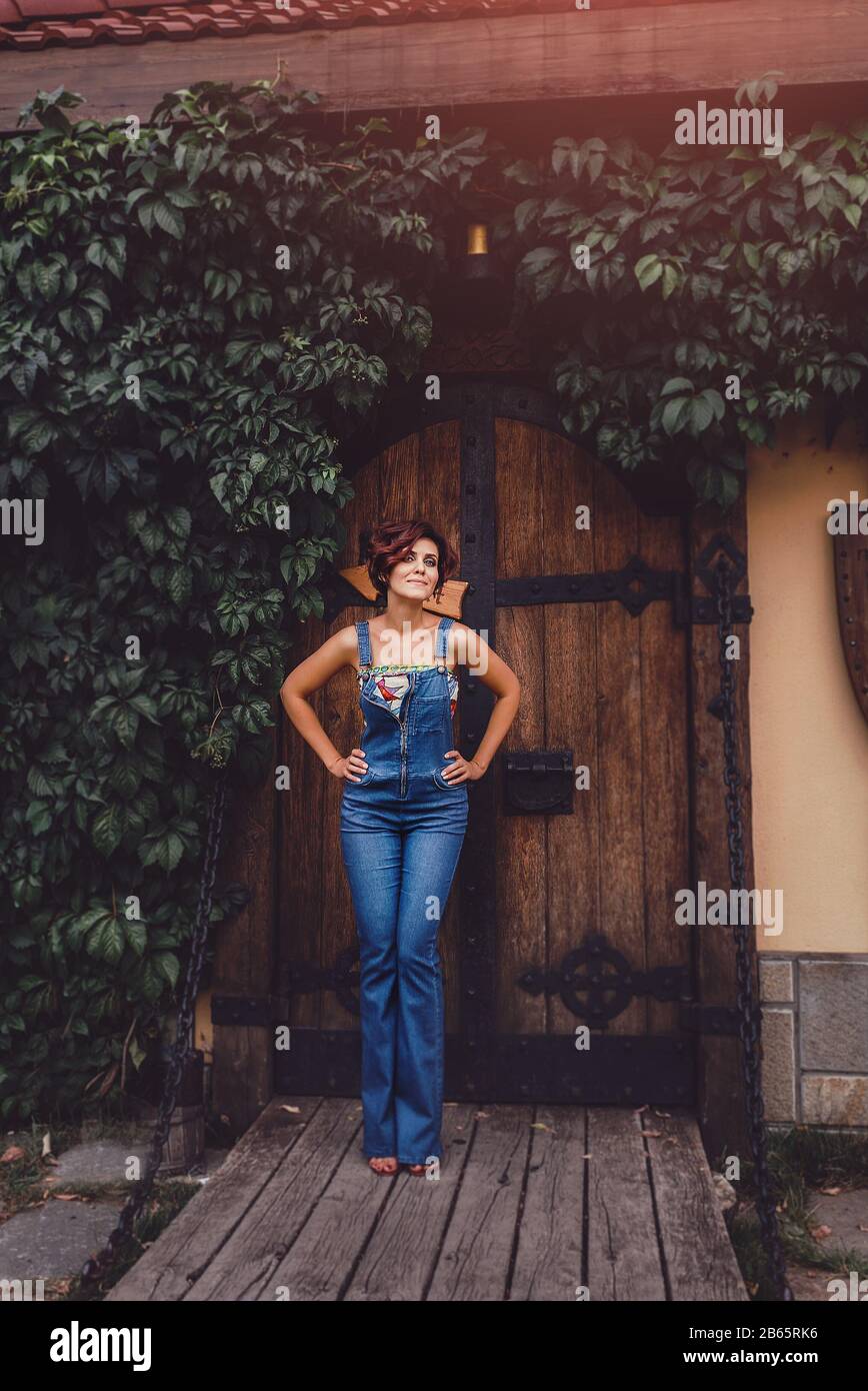 Porträt einer schönen Frau mit Denim-Jumpsuit, die gegen eine Efeue steht, die alte Holztür eines Schlosses umrahmt. Magisch gezont Stockfoto