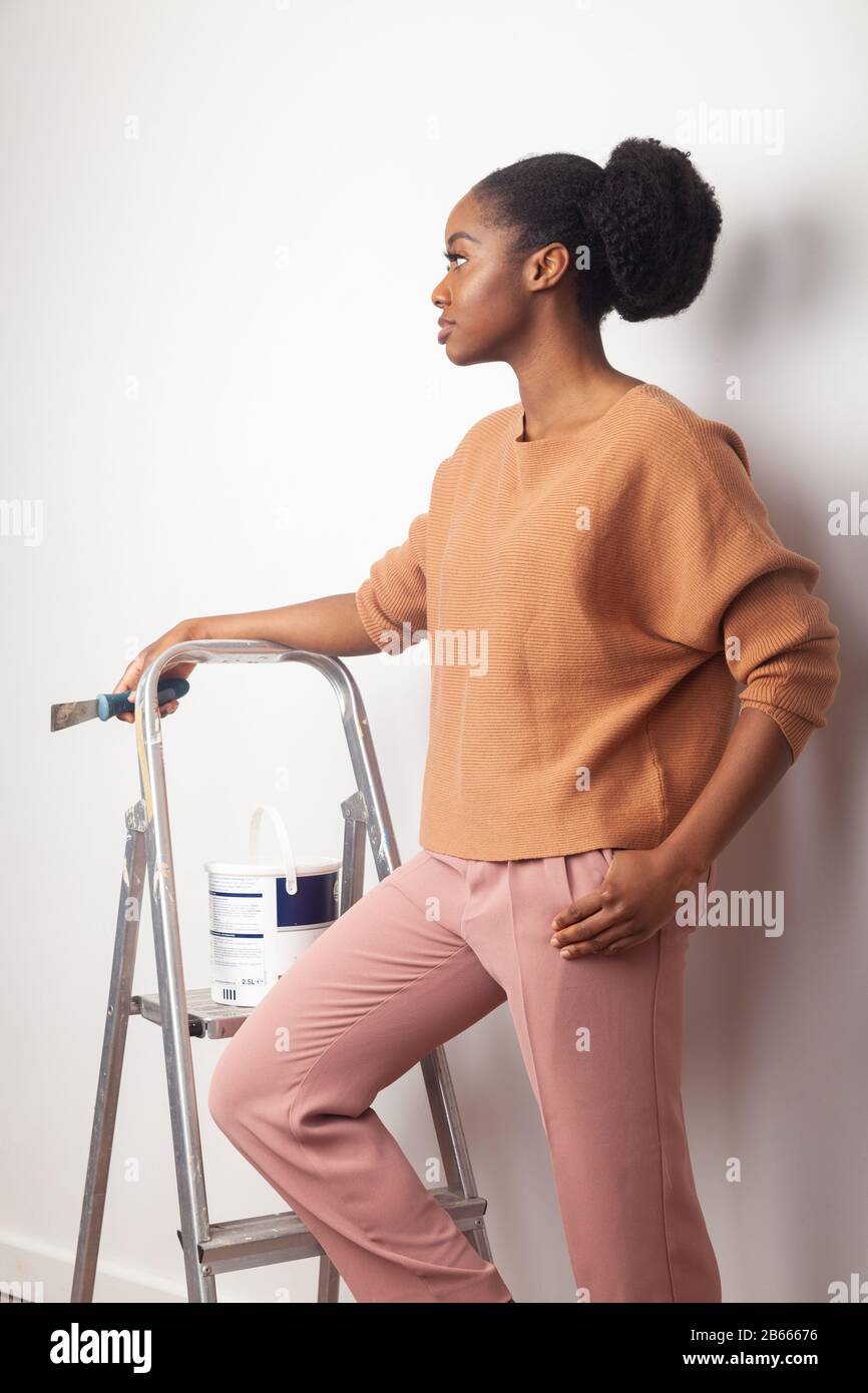 Eine schöne junge Frau afrikanischer Ethnie, die neben einer Stufenleiter steht und einen Tapetenkratzer in der Hand hält Stockfoto