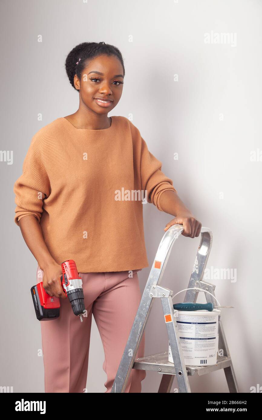Eine schöne junge Frau afrikanischer Ethnie, die neben einer Stufenleiter steht und eine elektrische Bohrmaschine hält Stockfoto