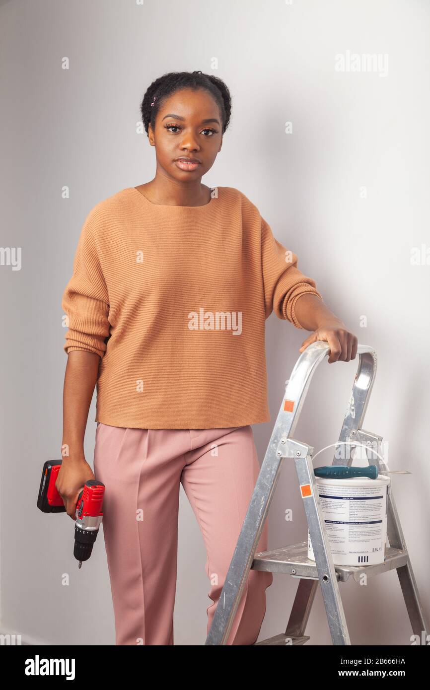 Eine schöne junge Frau afrikanischer Ethnie, die neben einer Stufenleiter steht und eine elektrische Bohrmaschine hält Stockfoto