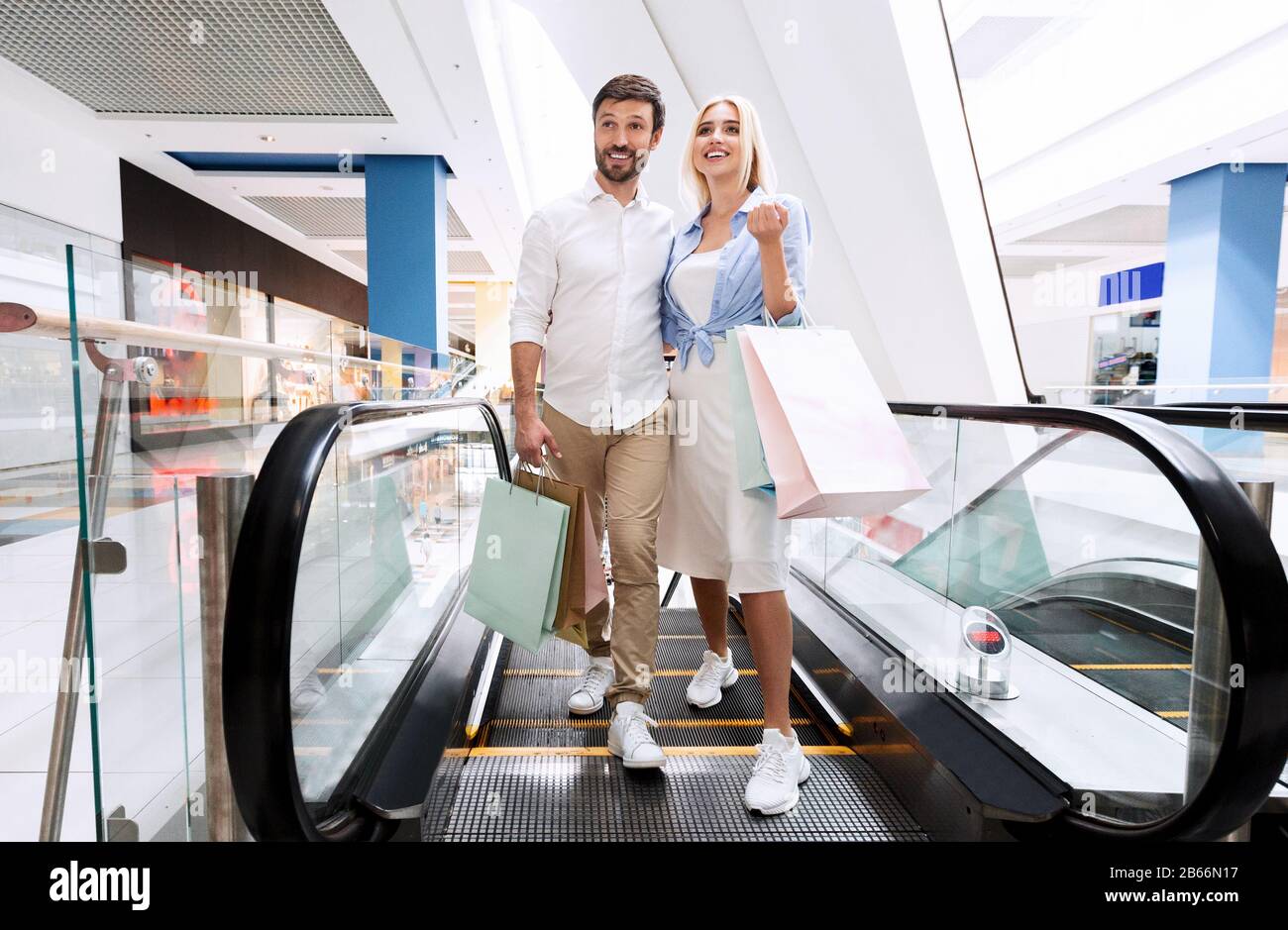Couple Holding Shopper Bags Getting Off Escalator Stairs In Mall Stockfoto