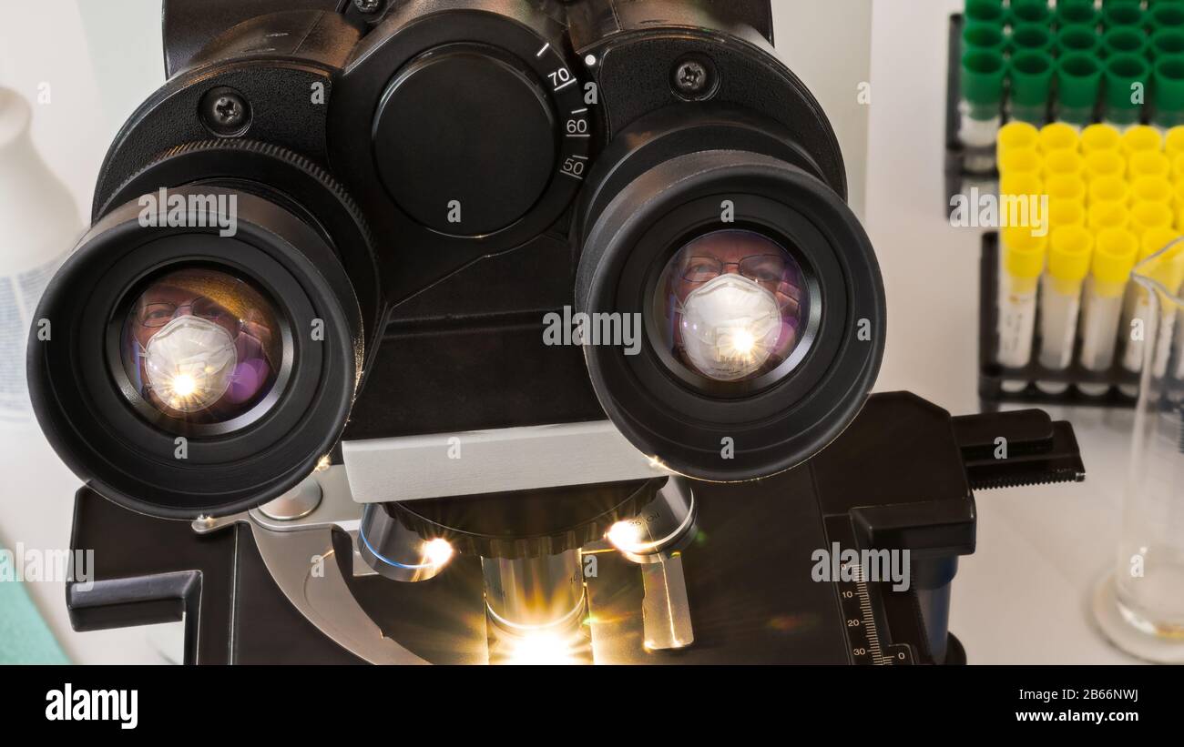 Laborlinse für medizinisches Mikroskop mit Spiegelung eines Manngesichts in weißer Operationsmaske. Optische Diagnosegeräte und Kunststoff-Reagenzgläser in Racks. Stockfoto