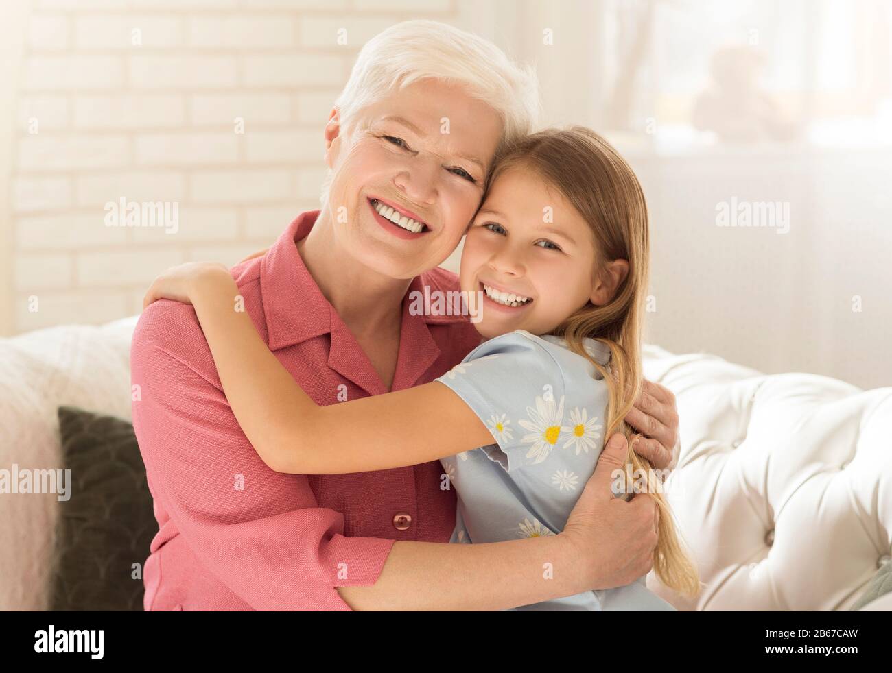 Fröhliche Granny mit Enkeltochter, die zu Hause auf dem Sofa umarmte Stockfoto