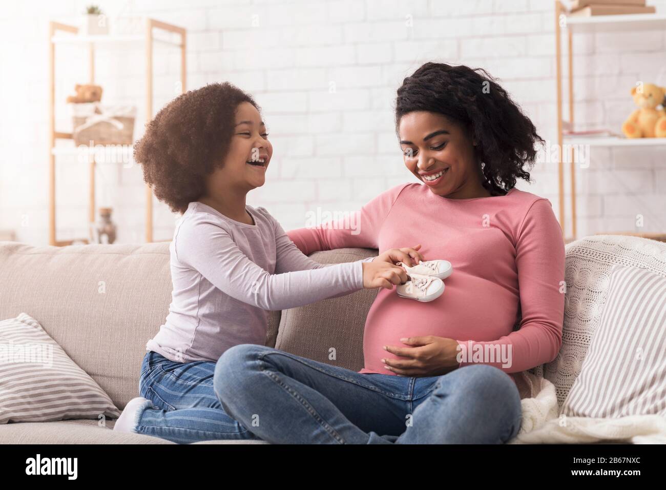 Adlige Tochter, Die Baby-Schuhe An Den Bauch Ihrer Schwangeren Afro Mum Legt Stockfoto