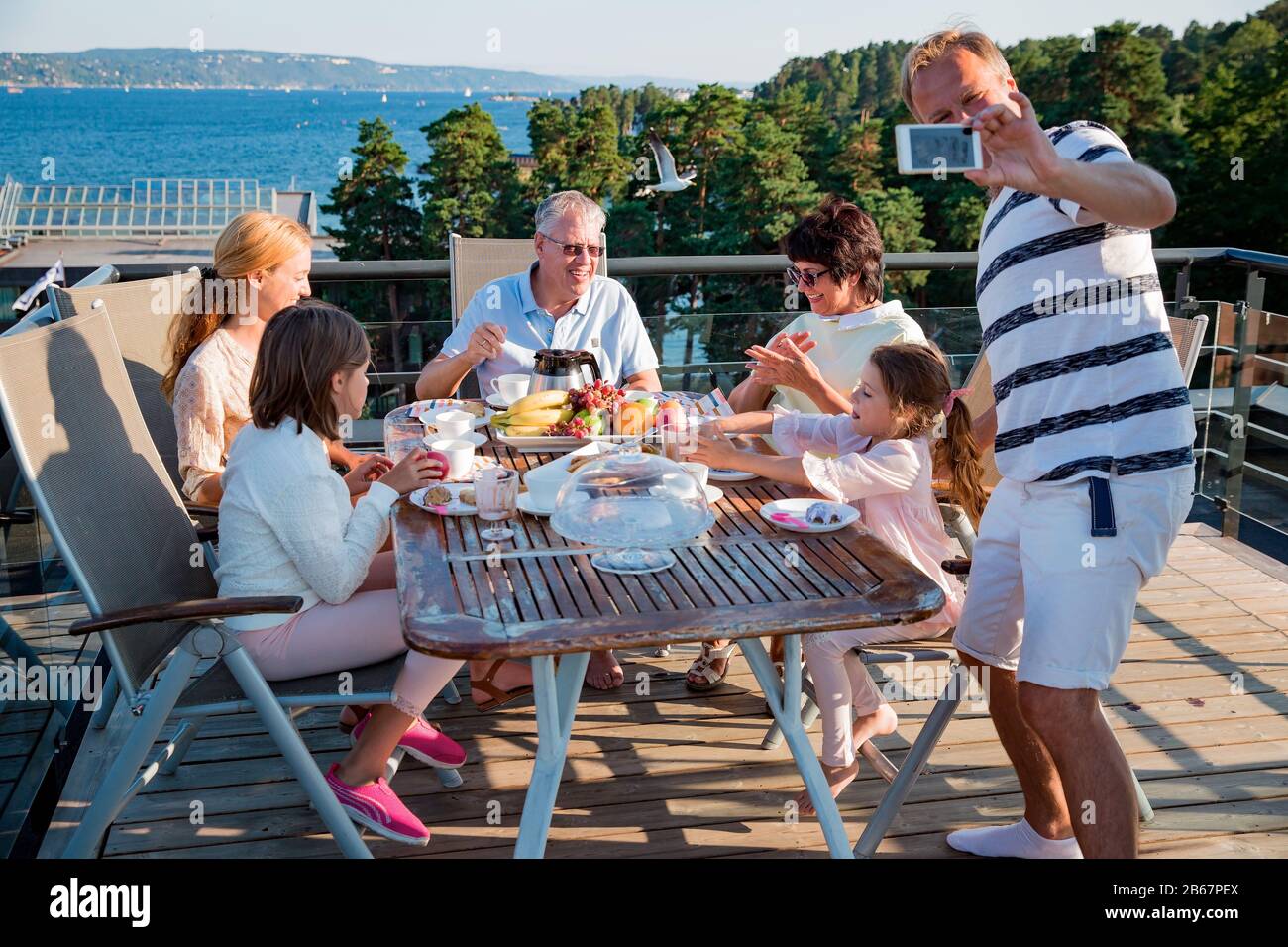 Große glückliche Familie, die zusammen auf der Terrasse frühstücken, am Tisch sitzen und Kaffee trinken. Vater nimmt selfie am Telefon. Schöne Aussicht Stockfoto