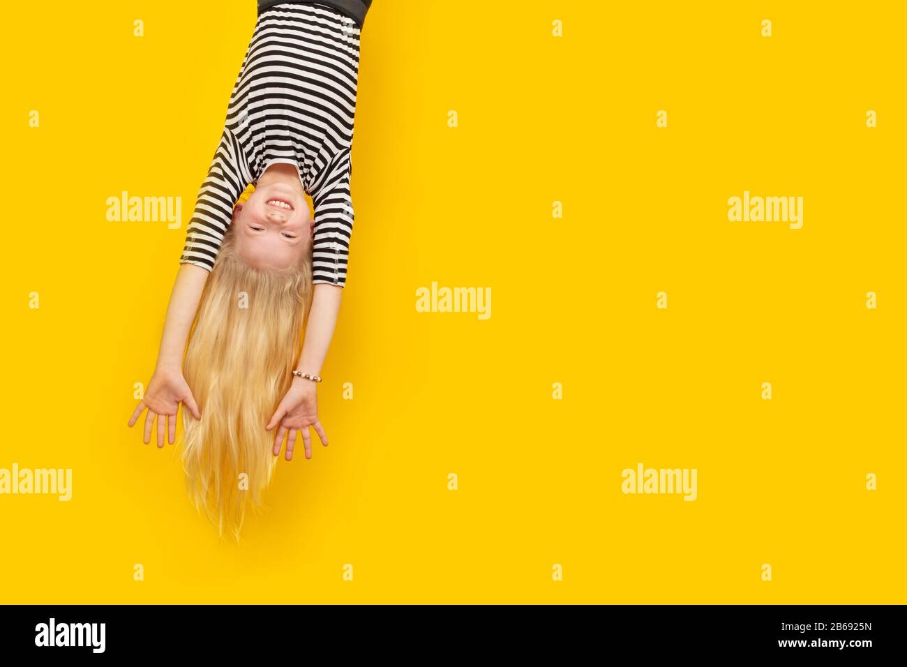 Aufgeregtes, verrücktes kleines blondes Mädchen, das glücklich auf dem Kopf hängt, übergibt sich über den isolierten gelben Studiohintergrund. Emotionen, Ausdruck. Platz für Text kopieren Stockfoto