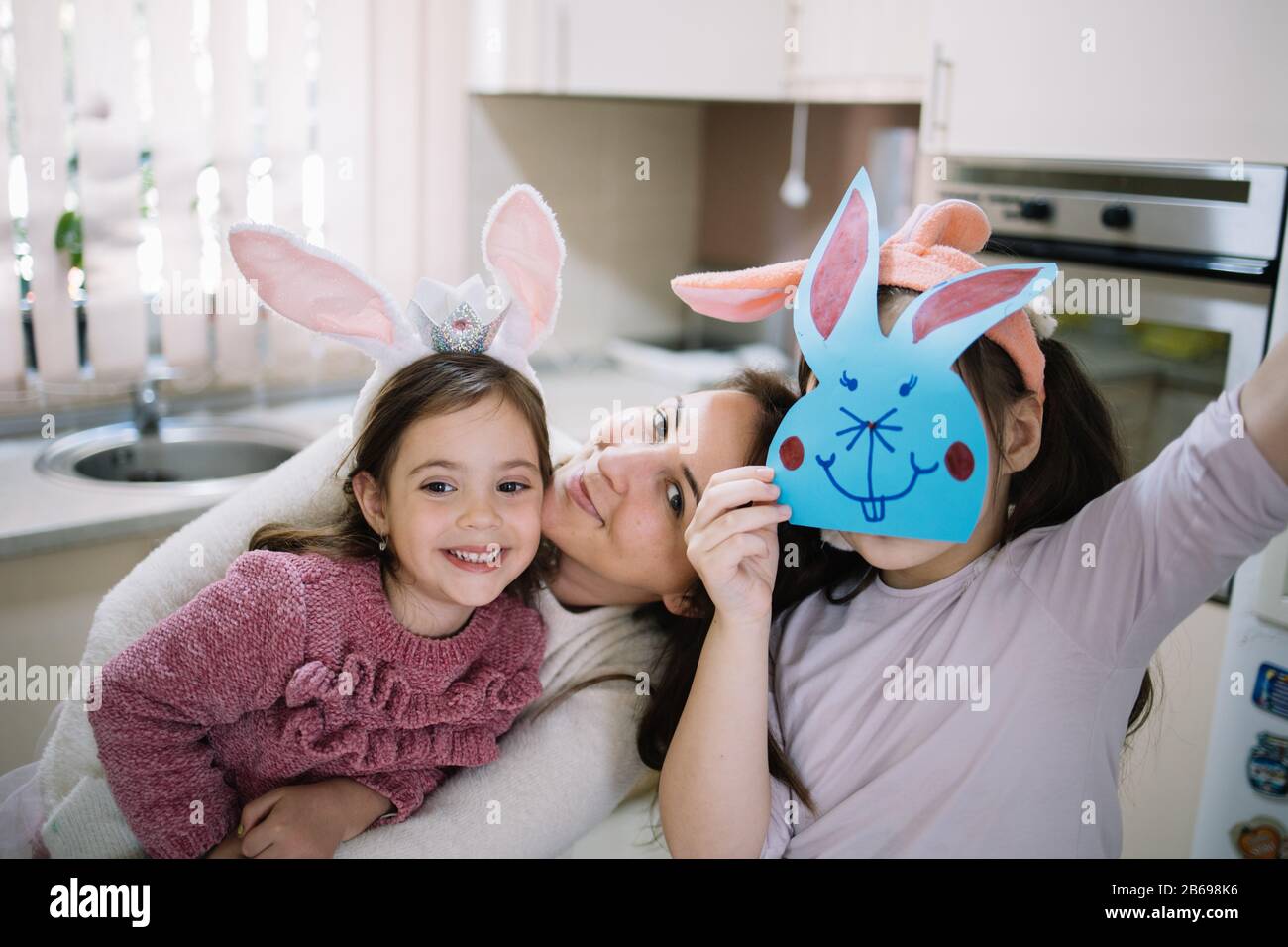 Lächelnde Mutter umarmte ihre Tochter mit Kaninchenohren. Fröhliche Mutter umarmt ihr Kind, während sie neben ihrer Tochter sitzt und mit Papierkaninchen auf dem Gesicht in der Sonne sitzt Stockfoto