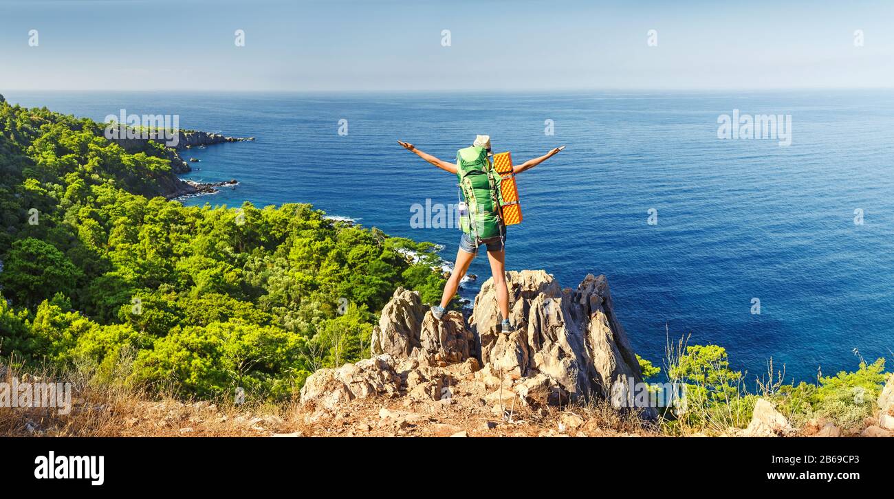 Eine junge glückliche Frau bei einer Wandertour mit einem Rucksack hob ihre Hände hoch und bewunderte den bunten Meerblick. Reise durch den Lykischen Weg in die Türkei Stockfoto