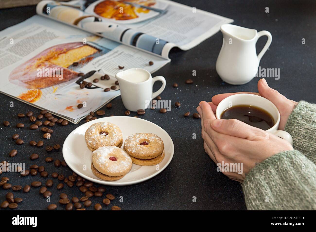 Tasse Kaffee in den Händen der Frau. Untertasse mit Plätzchen, gebratenen Kaffeebohnen und kulinarischem Magazin auf dunklem Tisch. Stockfoto