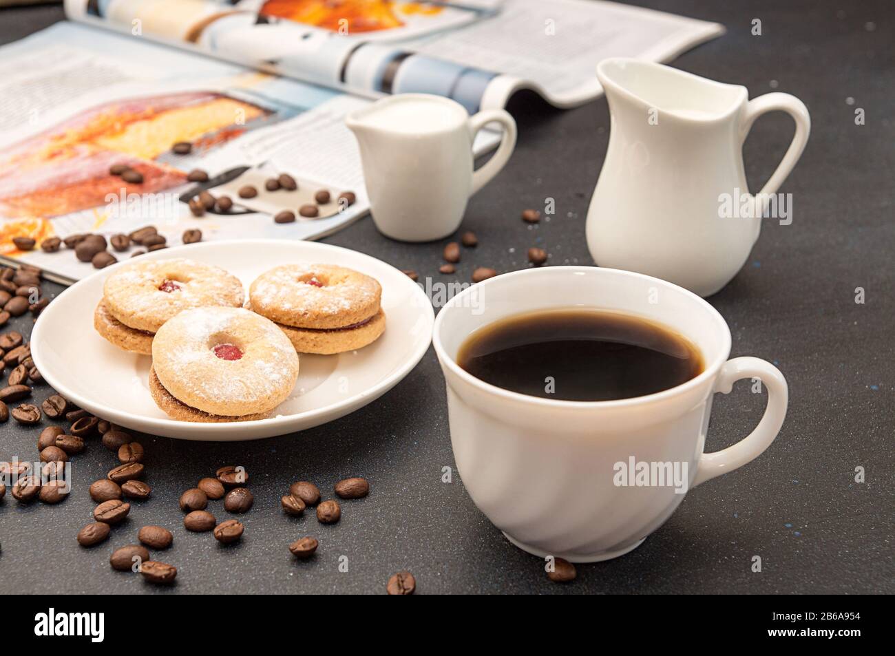 Tasse Kaffee, Untertasse mit Plätzchen, Milchkanne, gebratene Kaffeebohnen und kulinarisches Magazin auf dunklem Tisch. Stockfoto