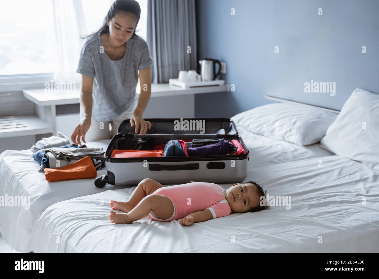 Baby liegt auf dem Bett, wenn seine Mutter seine Kleidung in einen Koffer legt, bevor sie in den Urlaub geht Stockfoto
