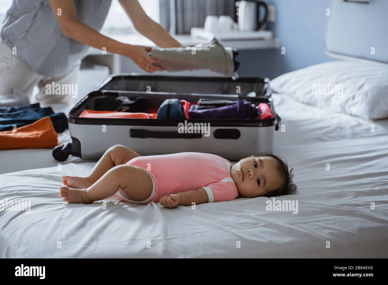 Süßes Baby, das auf dem Bett neben einem Koffer liegt und mit Kleidung gefüllt ist, die für eine Urlaubsreise vorbereitet ist Stockfoto