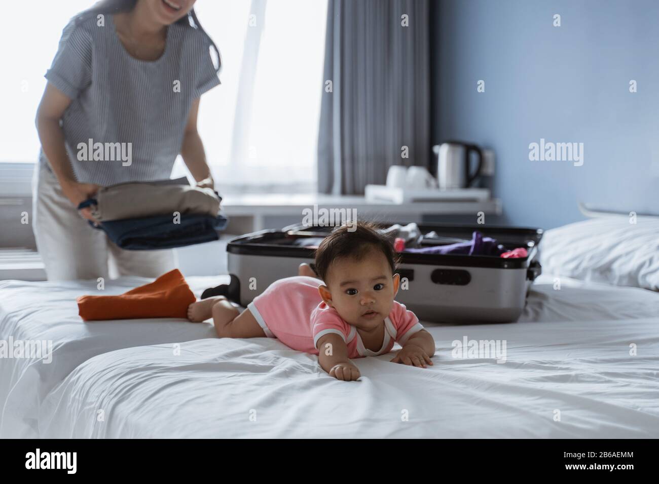 Baby, das auf dem Bett neben einem Koffer liegt und mit Kleidung gefüllt ist, die für eine Urlaubsreise vorbereitet ist Stockfoto