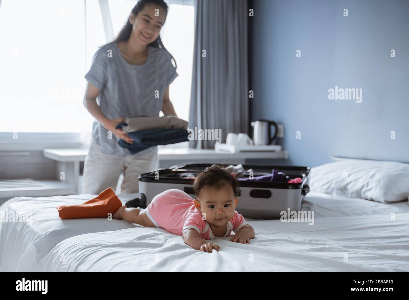 Süßes Baby liegt auf dem Bett, wenn seine Mutter seine Kleidung in einen Koffer legt, bevor sie in den Urlaub geht Stockfoto