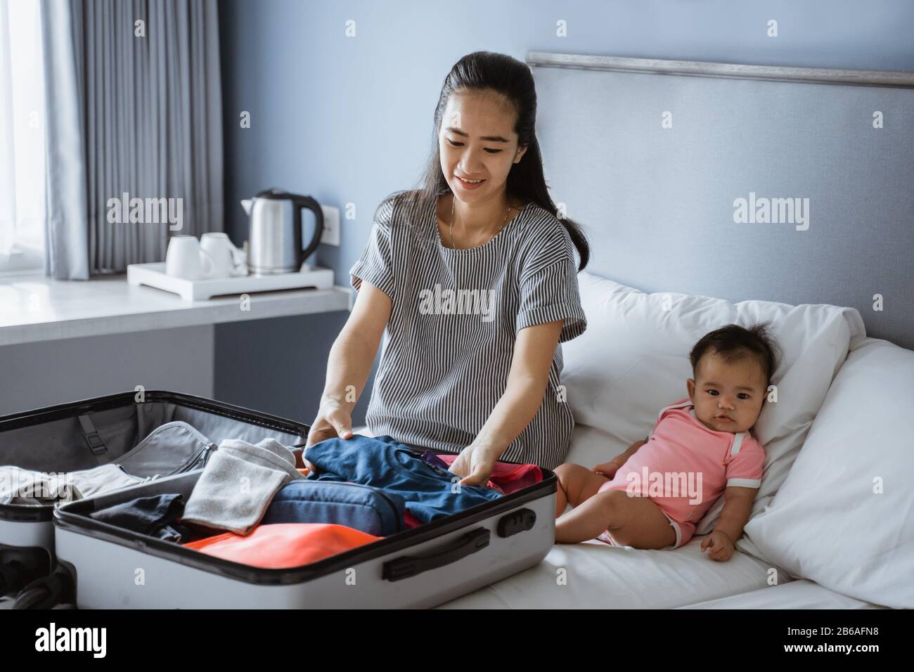 Baby sitzt neben der Mutter und bereitet Kleidung zum Einlegen in den Koffer vor Stockfoto