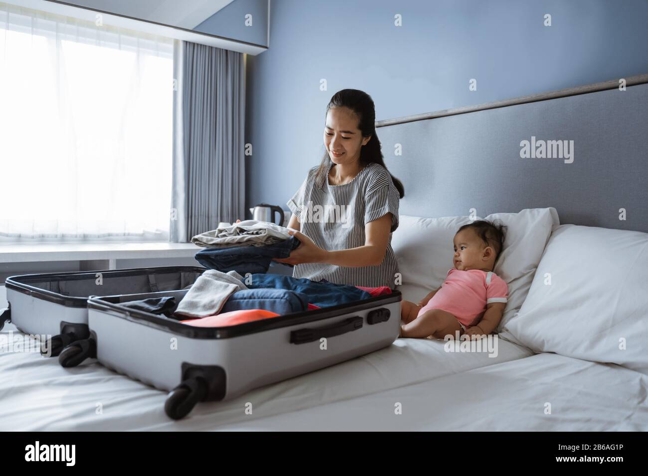 Baby sitzt neben der Mutter und bereitet Kleidung zum Einlegen in den Koffer vor Stockfoto