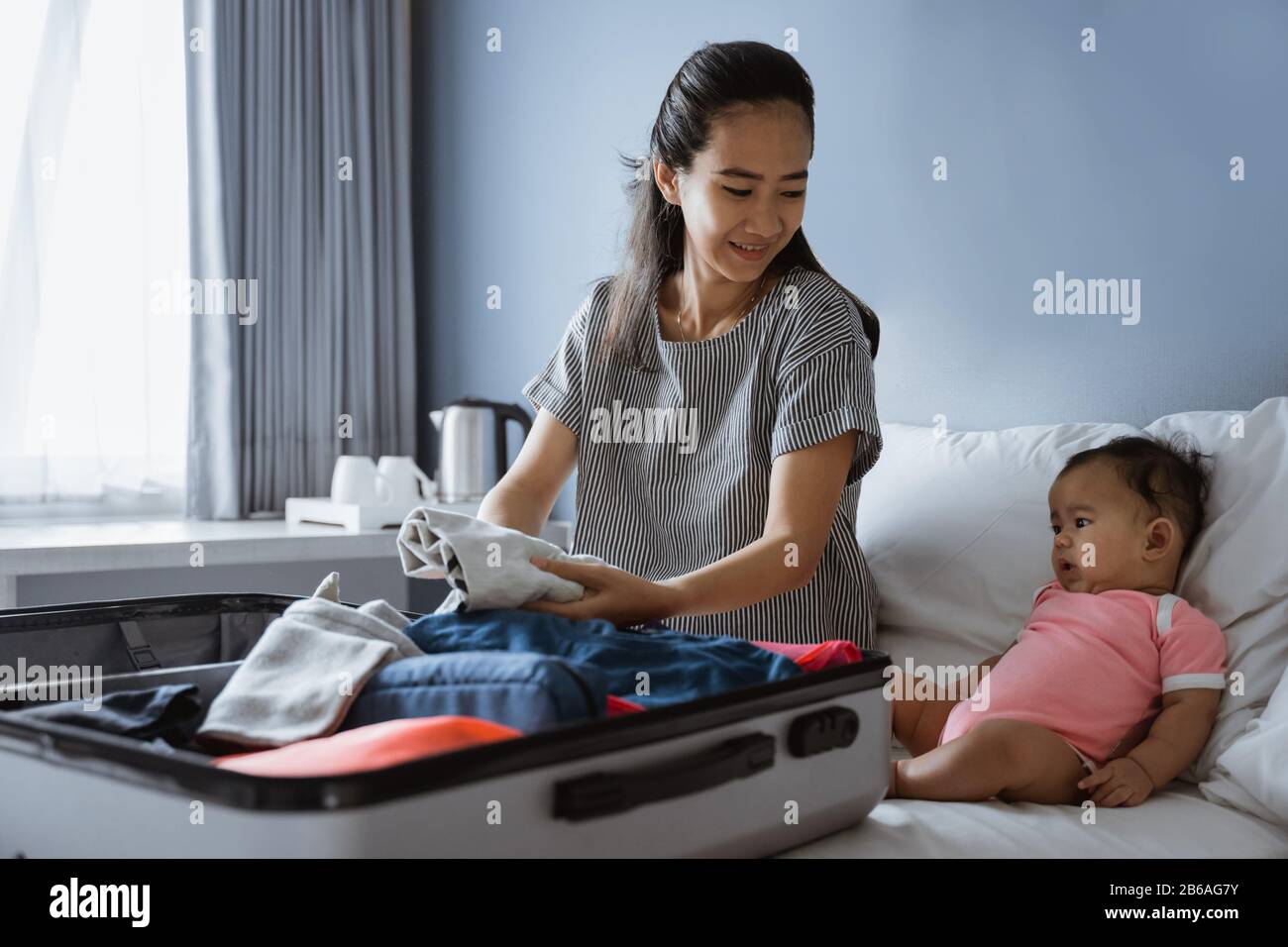 Süßes Baby sitzt neben der Mutter und bereitet Kleidung zum Einlegen in den Koffer vor Stockfoto