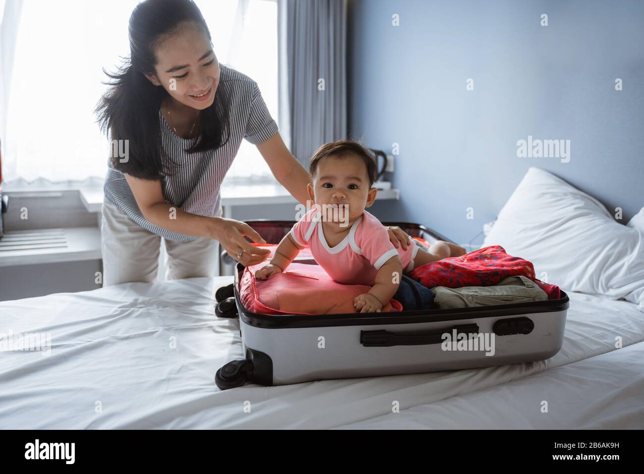 Mama lächelt, als sie ein niedliches Baby in einem offenen Koffer mit  Kleidung auf dem Bett liegen hält Stockfotografie - Alamy