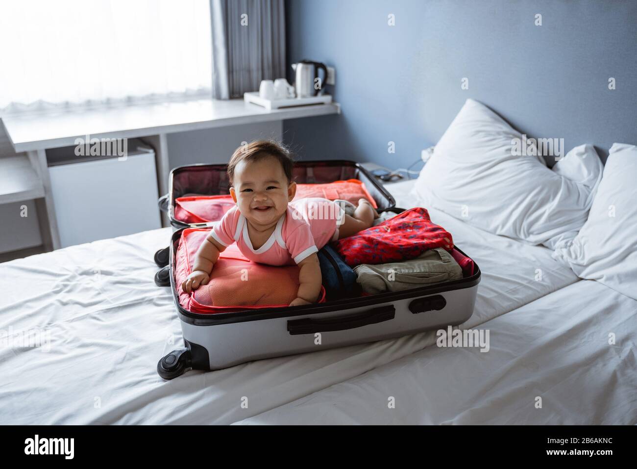 Baby lacht auf dem Bauch in einem offenen Koffer, der mit Kleidung gefüllt ist Stockfoto