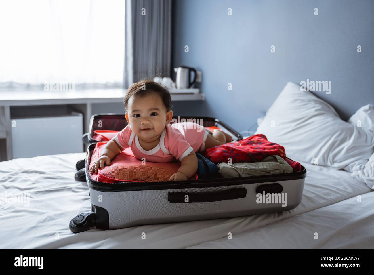 Baby lächelt auf dem Bauch in einem offenen Koffer, der mit Kleidung gefüllt ist Stockfoto