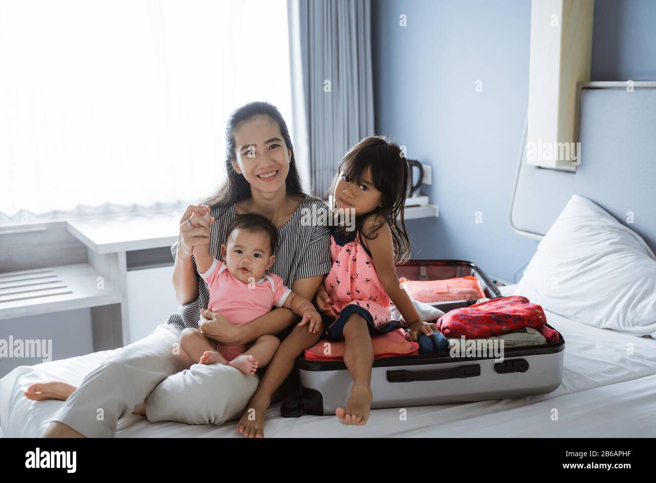 Mutter und ihre beiden Kinder lächeln, nachdem sie einen Koffer für den Urlaub vorbereitet haben, auf dem Bett sitzend Stockfoto
