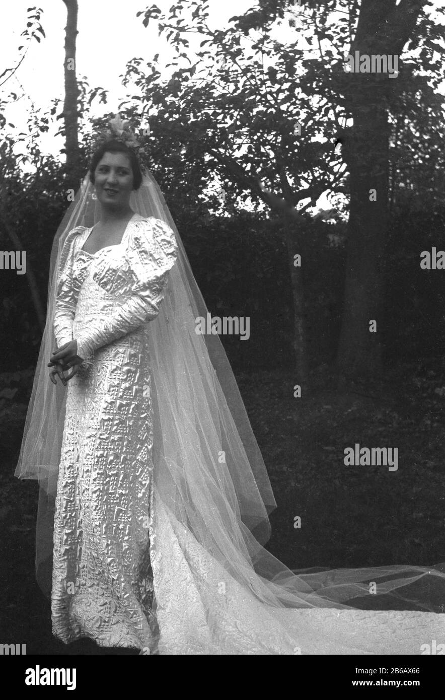 Dreißiger Jahre, historisch, Brautpaar mit langem Schleier, der draußen in einem ungewöhnlichen, glänzenden Hochzeitskleid steht, England, Großbritannien. Stockfoto