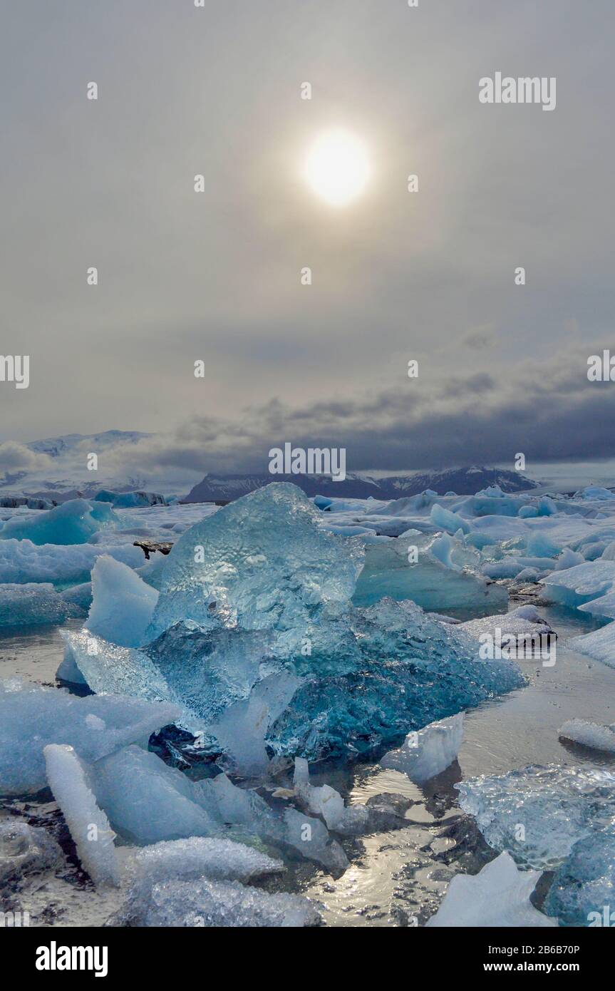 Glitzernde blaue Eisberge schweben an einem bewölkten Tag in der Lagune des Jokulsarlon-Ozeans mit den Bergen im Hintergrund und der Sonne darüber - Island Stockfoto