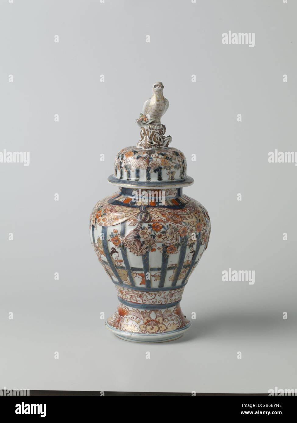 Balustervase mit Abdeckung für Menschen in einem inneren Heckgitter, prunusbäumen und Blumenrinnen Balustervormige Dekselpot van Porselein traf een spreidende voet, beschilderd in onderglazuur blauw en op het glazuur rood, zwart en goud. OP de Wand twee keer twee mensen zittend in een interieur achter gemodelleerd en beschilderd traliewerk. IT traliewerk is versierd met gemodelleerde en gedecoreerde prunusbomen en banden met bloemranken. Onder het traliewerk een Band met bloemranken. Rond de voet een Band met lotusranken. OP de hals drie Cartuches met lotusranken. De deksel traf Dezelfde decorati Stockfoto
