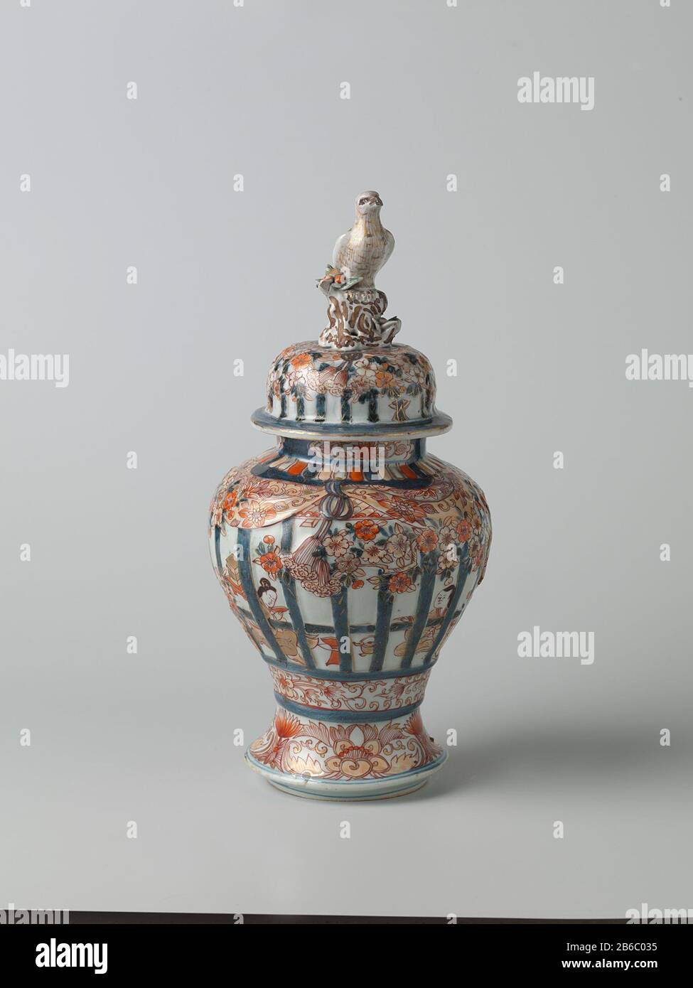Balustervase mit Abdeckung für Menschen in einem inneren Heckgitter, prunusbäumen und Blumenrinnen Balustervormige Dekselpot van Porselein traf een spreidende voet, beschilderd in onderglazuur blauw en op het glazuur rood, zwart en goud. OP de Wand twee keer twee mensen zittend in een interieur achter gemodelleerd en beschilderd traliewerk. IT traliewerk is versierd met gemodelleerde en gedecoreerde prunusbomen en banden met bloemranken. Onder het traliewerk een Band met bloemranken. Rond de voet een Band met lotusranken. OP de hals drie Cartuches met lotusranken. Imari. Hersteller: Pottenbak Stockfoto