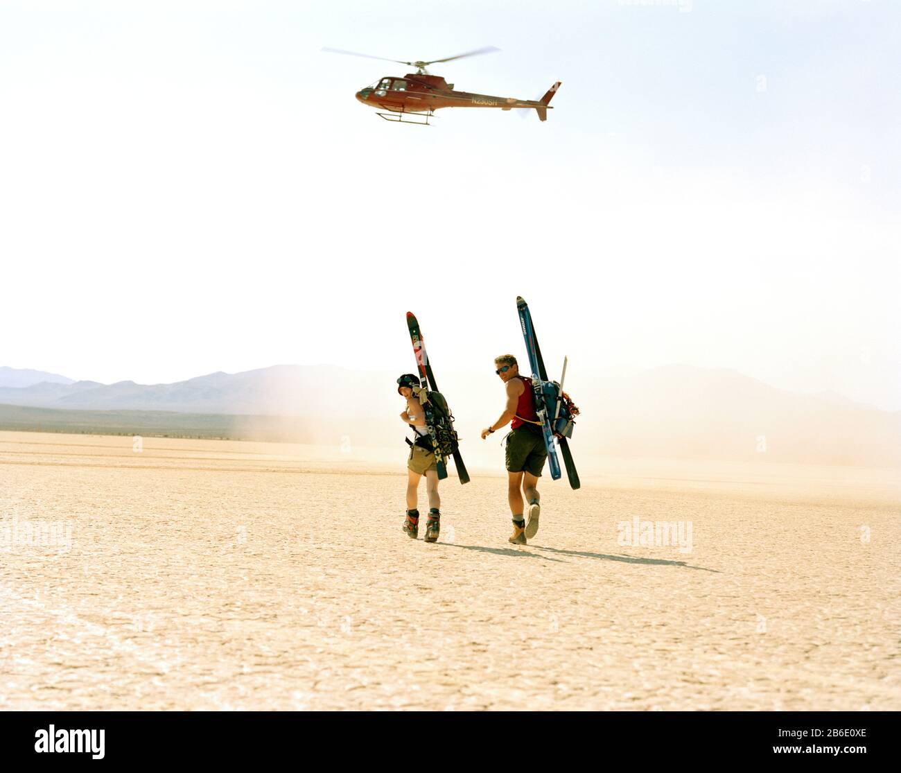 Porträt eines abenteuerlustigen jungen Paares, das Sportausrüstung auf dem Rücken unter dem schwebenden Hubschrauber trägt, während er durch eine Wüste spaziert. Stockfoto