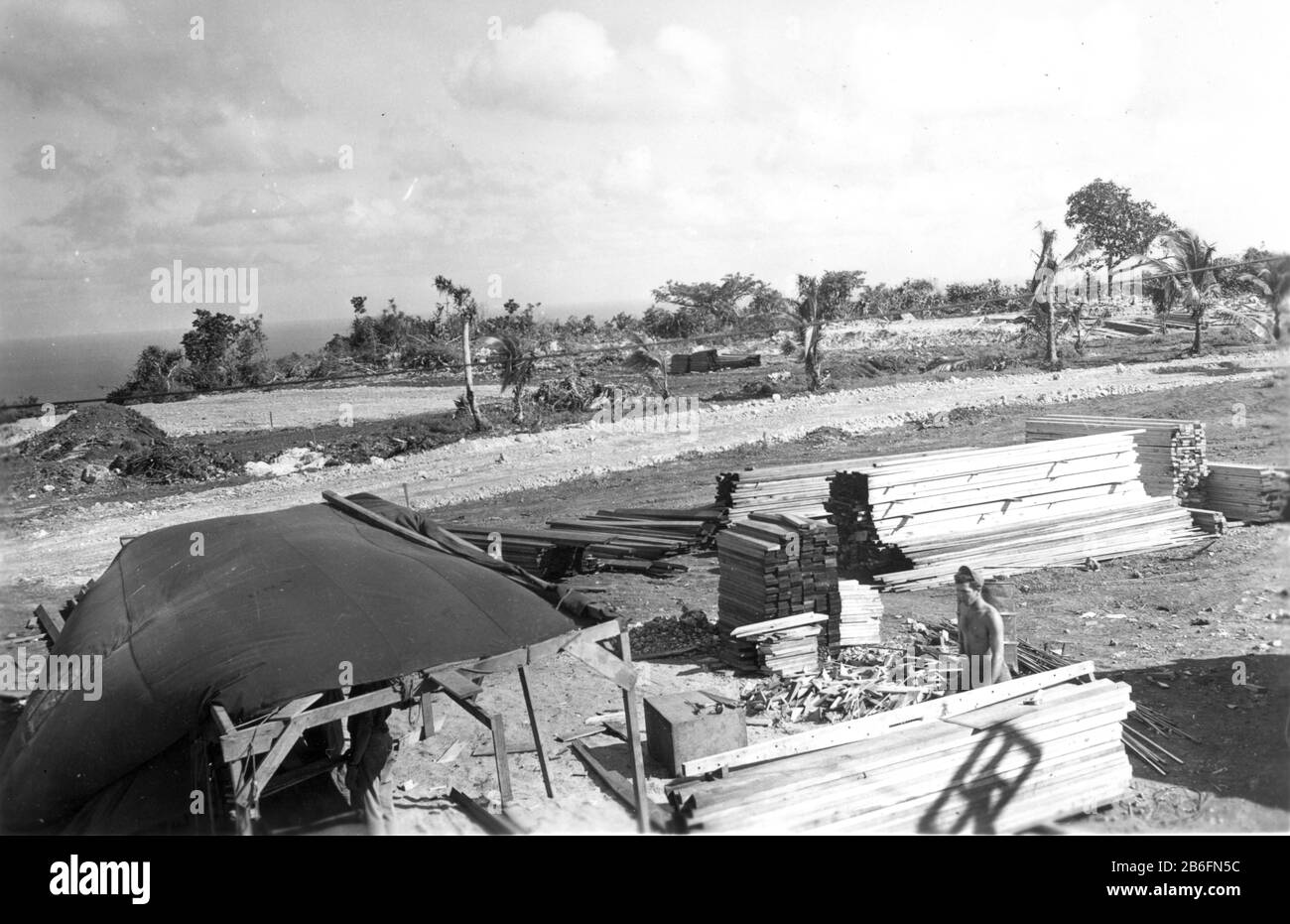 Seabees on Guam, # 3, Nov. 1944. Ein Mann im Lean-to und ein anderer arbeiten im Montagebereich für kommende Gebäude. Beachten Sie die Werkzeuge auf einem Kasten rechts neben dem Lean-to. Dieses Foto zeigt eine neu ausrauchende Schotterstraße vor den zerbrochenen Bäumen hinter den Magen- und Holzhaufen. Das zeigt auch zwei weiß-weiße Bodenbereiche hinter der Straße, die für kommende Gebäude abgestuft wurden. Diese Seabees arbeiten an der Errichtung von Gebäuden für die Gebäude der CINCPAC-Offiziere. Um meine zugehörigen Vintage-Bilder zu sehen, suchen Sie nach Prestor Vintage WW II Stockfoto