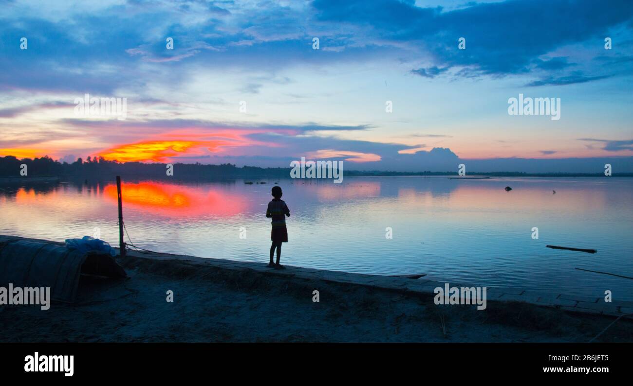 Ein Junge steht allein am Ufer eines Flusses im Abendlicht im Norden Bangladeschs. Die Schönheit des Augenblicks, der den Jungen wie einen Zorn anschrie. Stockfoto