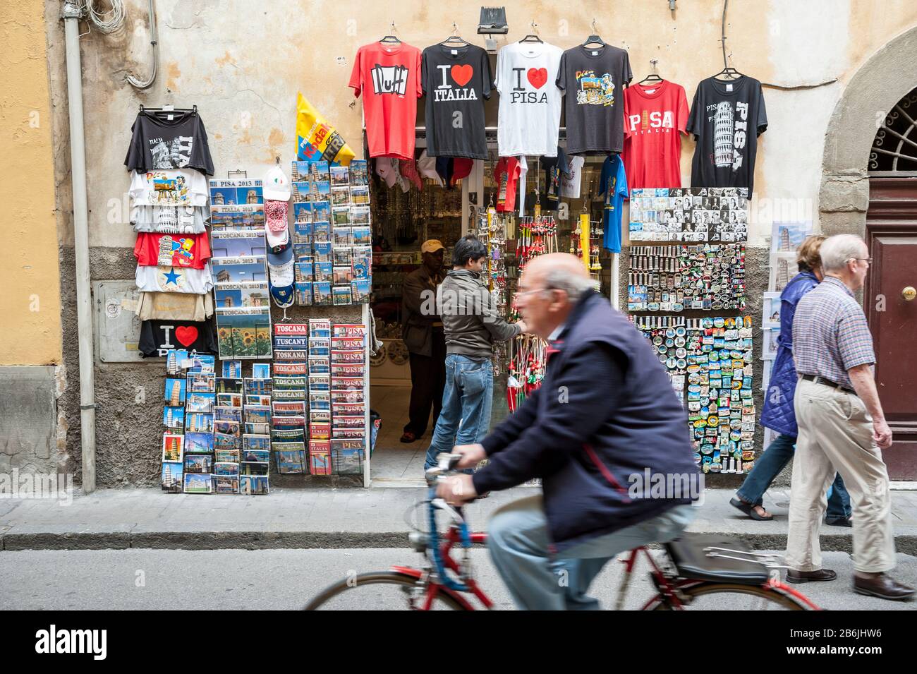 PISA, ITALIEN - 23. MAI 2012: Ein italienischer Radfahrer passiert einen Souvenirstand, der T-Shirts und Trinkets auf einer Straße zum Schiefen Turm verkauft. Stockfoto