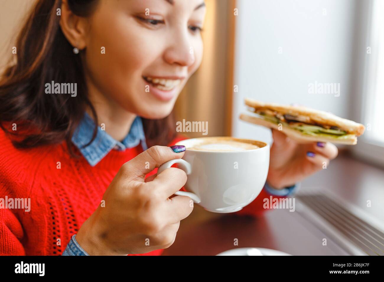 Junge Studentin isst ein Sandwich und trinkt Kaffee in einem Café in der Nähe des Fensters, Büro-Dinner-Pausenkonzept Stockfoto