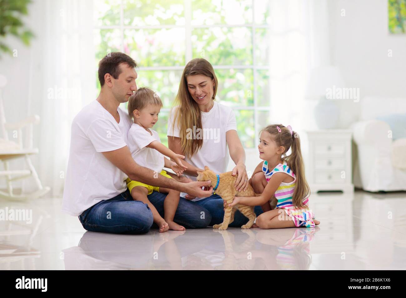 Glückliche junge Familie mit Kindern zu Hause. Vater, Mutter, Tochter und Sohn spielen im Wohnzimmer auf dem Boden. Mutter, Vater, kleiner Junge und Mädchen, die ein Haustier halten Stockfoto