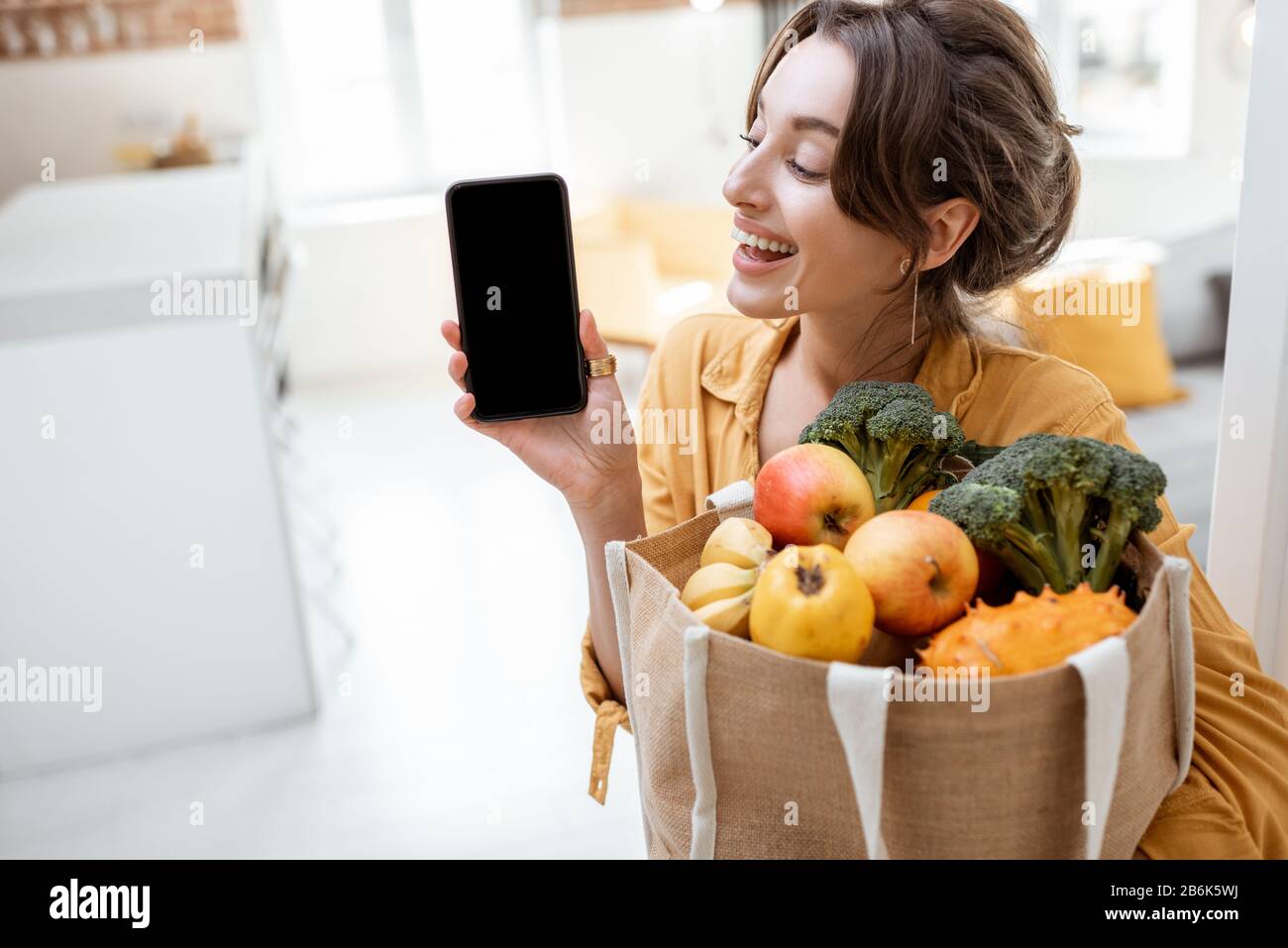 Portrait einer jungen und fröhlichen Frau mit Smartphone und Einkaufstasche voller frisches Obst und Gemüse zu Hause. Telefon mit leerem Bildschirm zum Kopieren des Einfügevorgangs Stockfoto