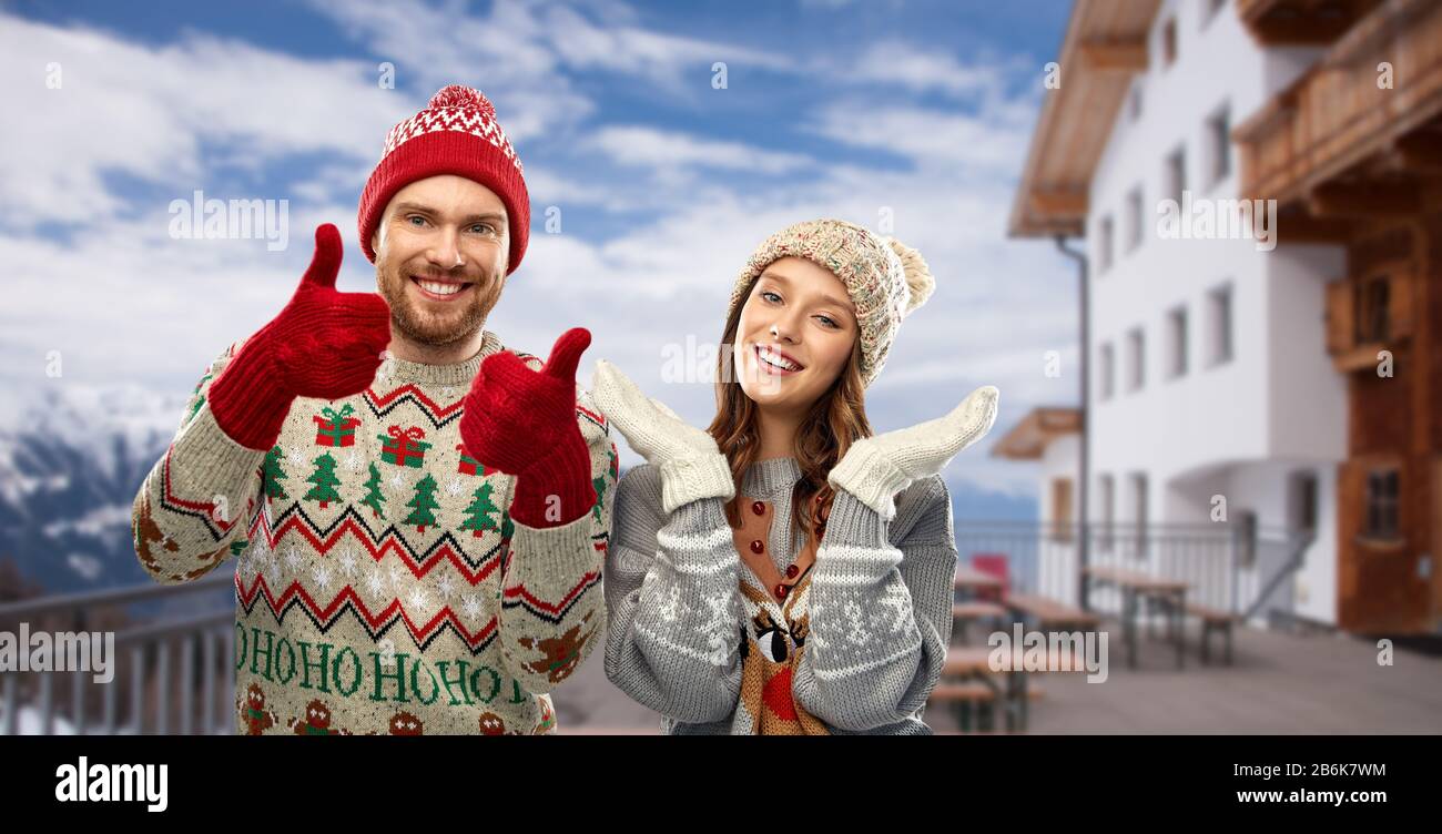 Weihnachten, Winterkleidung und Weihnachtskonzept - glückliches Paar in hässlichen Pullovern, Strickmützen und Fäusten mit Daumen nach oben über dem Skigebiet in österreich Stockfoto