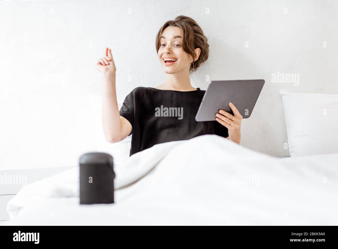 Junge Frau, die mit einem digitalen Tablet und einem intelligenten Lautsprecher im Bett liegt und die Heimgeräte mit Sprachbefehlen steuert. Konzept für ein gepflegtes Zuhause und Entspannung zu Hause Stockfoto
