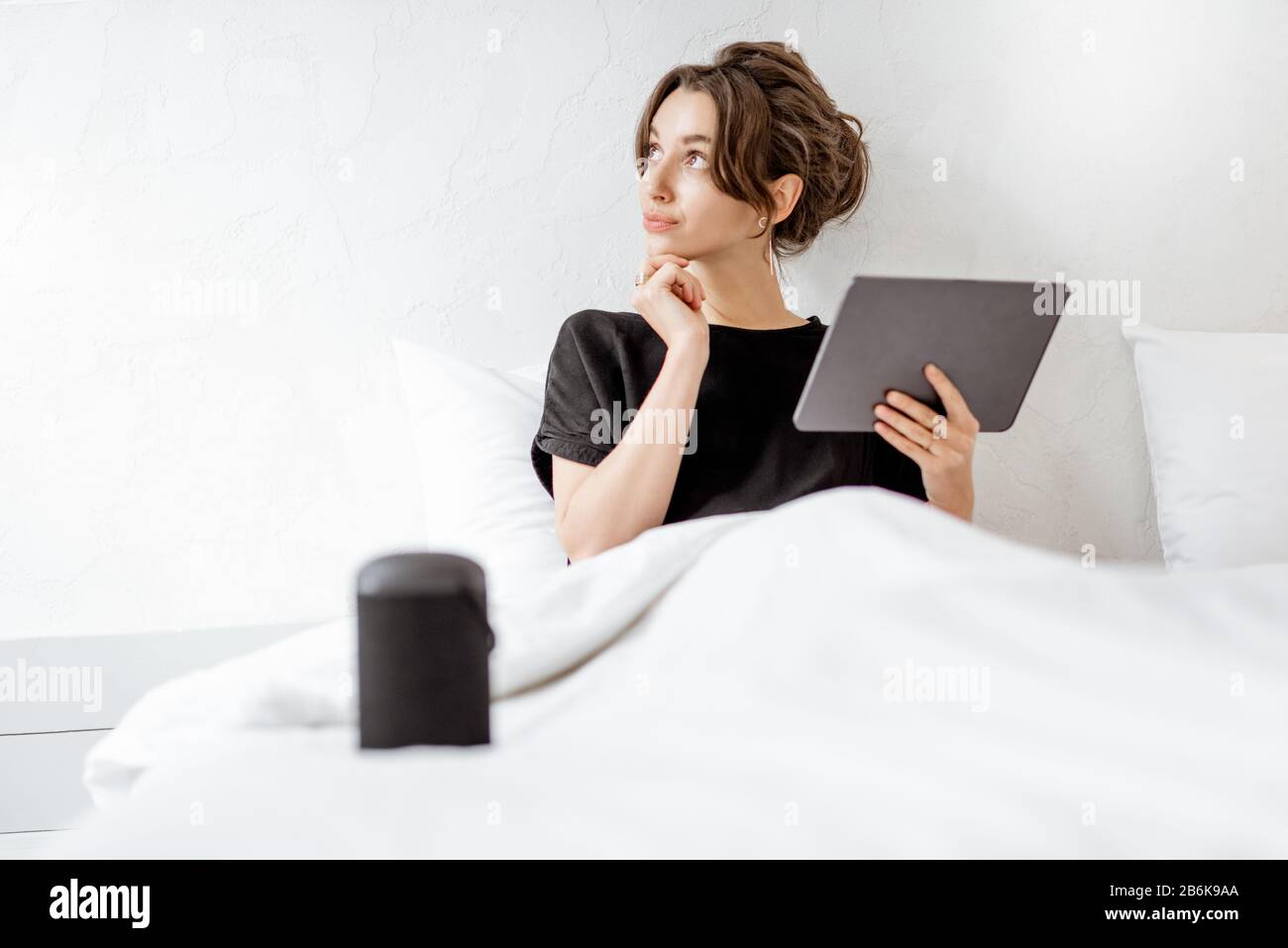 Junge Frau, die mit einem digitalen Tablet und einem intelligenten Lautsprecher im Bett liegt und die Heimgeräte mit Sprachbefehlen steuert. Konzept für ein gepflegtes Zuhause und Entspannung zu Hause Stockfoto