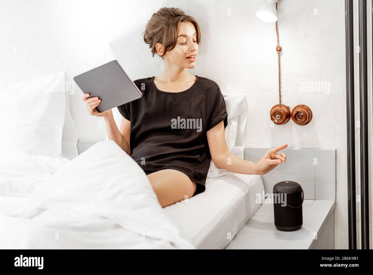 Junge Frau, die mit einem digitalen Tablet und einem intelligenten Lautsprecher im Bett liegt und die Heimgeräte mit Sprachbefehlen steuert. Konzept für ein gepflegtes Zuhause und Entspannung zu Hause Stockfoto