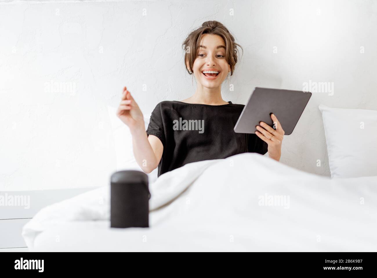 Junge Frau, die mit einem digitalen Tablet und einem intelligenten Lautsprecher im Bett liegt und die Heimgeräte mit Sprachbefehlen steuert. Konzept für ein gepflegtes Zuhause und Entspannung zu Hause Stockfoto