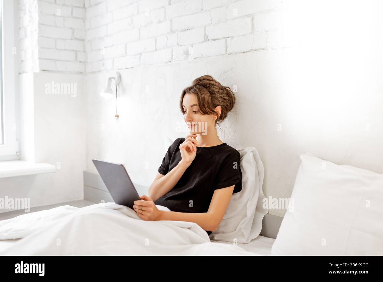 Junge Frau, die digitale Tablette verwendet, während sie entspannt im Bett eines weißen Bedrroms sitzt. Konzept einer Freizeit mit mobilen Funkgeräten zu Hause Stockfoto