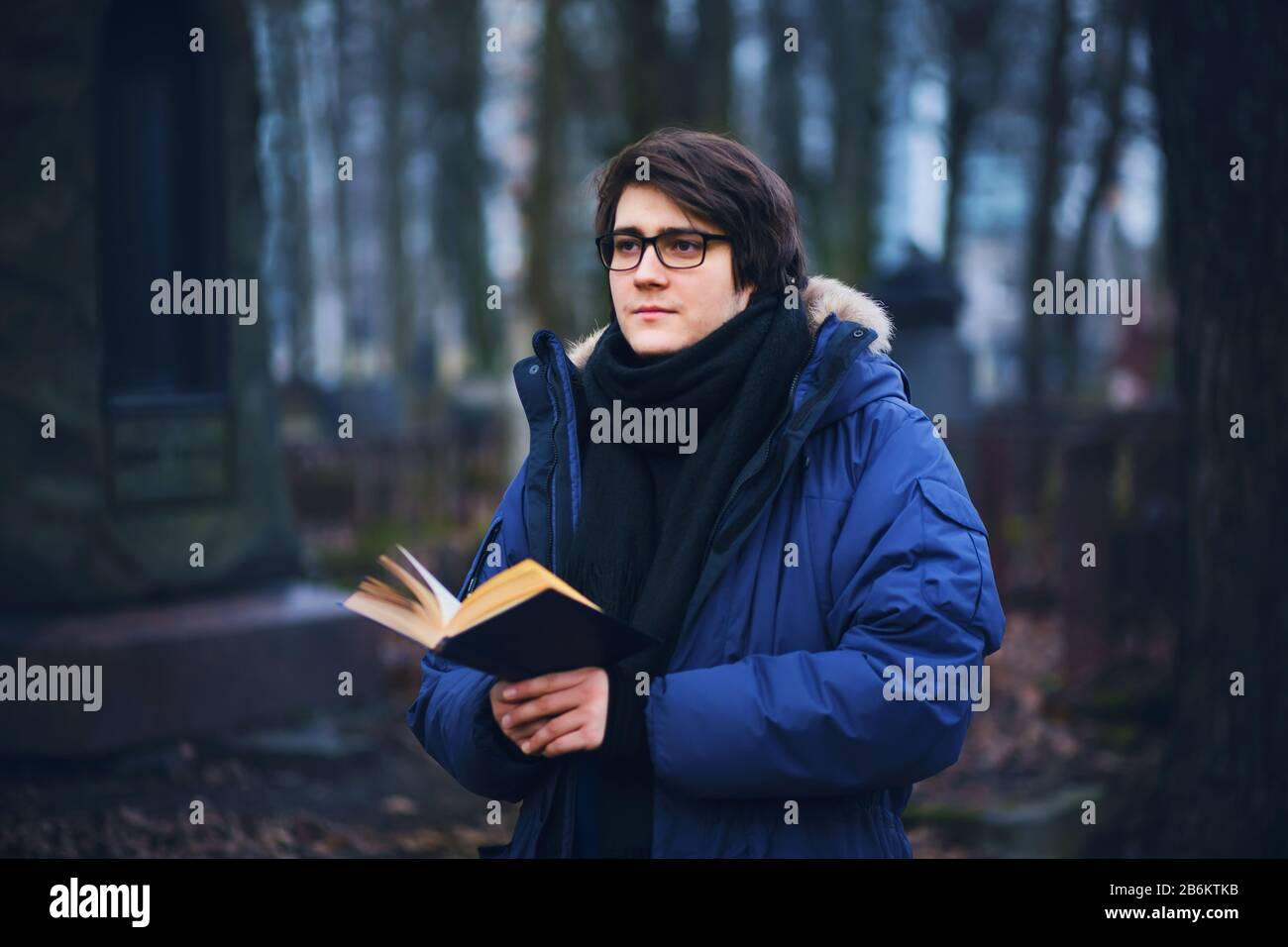 Ein junger ernsthafter Student in einer blauen Jacke im kalten Herbst steht im Park an der Gedenkstätte mit einem Buch und ehrt die Erinnerung in der dunklen Zeit von t Stockfoto