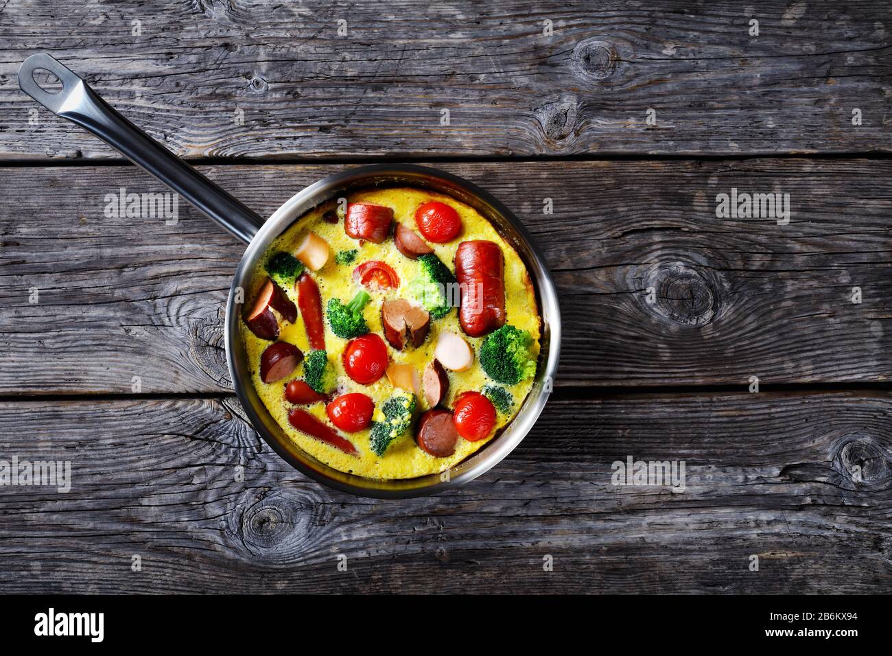 Gebackene Omelette oder Frittata mit Würstchen, Brokkoli, Kirschtomaten, geschmolzenem Käse im Skillet auf einem Holztisch im Landhausstil, horizontale Sicht von oben, fl Stockfoto