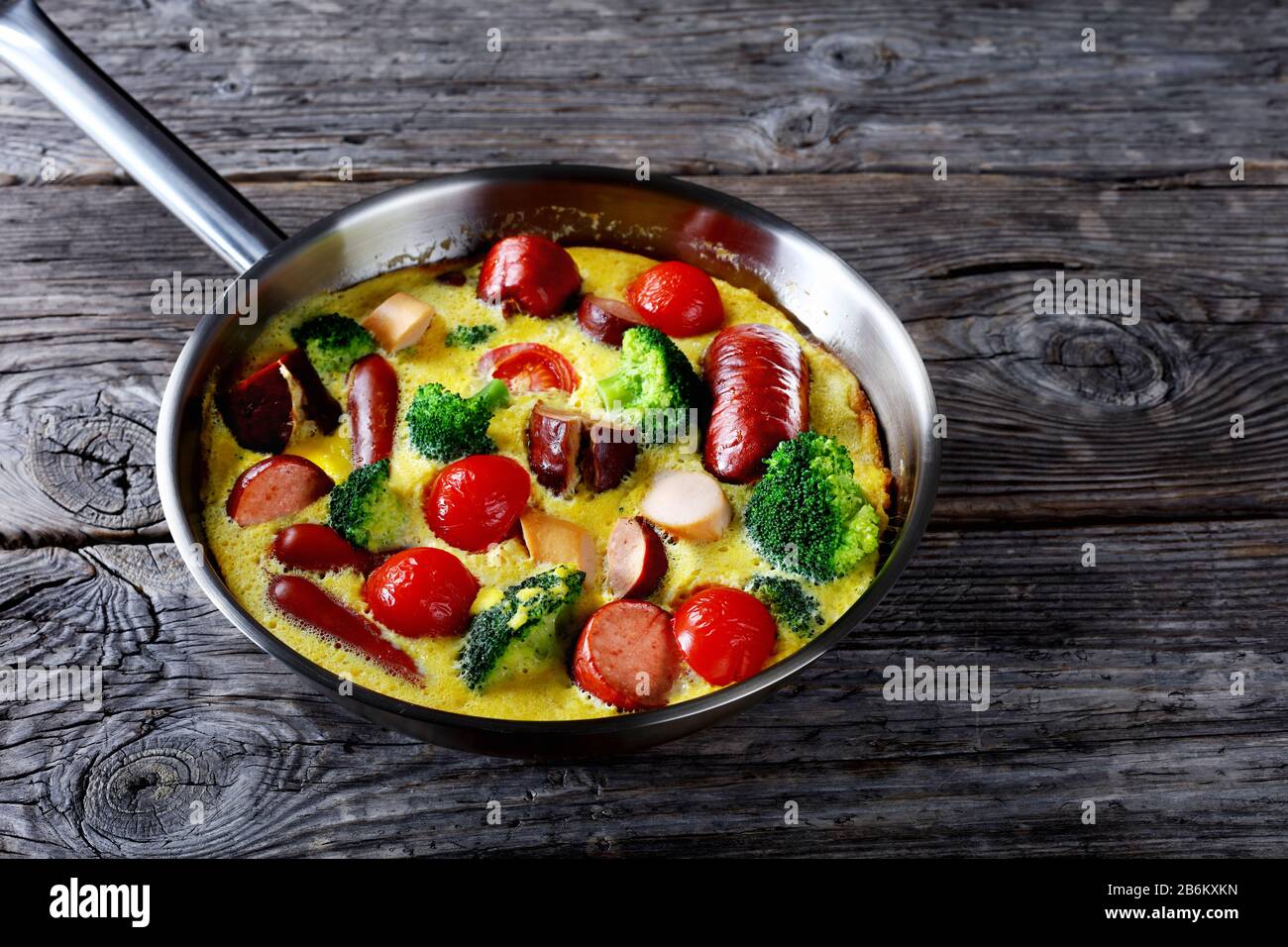 Gebackene Omelette oder Frittata mit Würstchen, Brokkoli, Kirschtomaten, geschmolzenem Käse in einem Skillet auf einem Holztisch im Landhausstil, horizontale Ansicht von oben Stockfoto