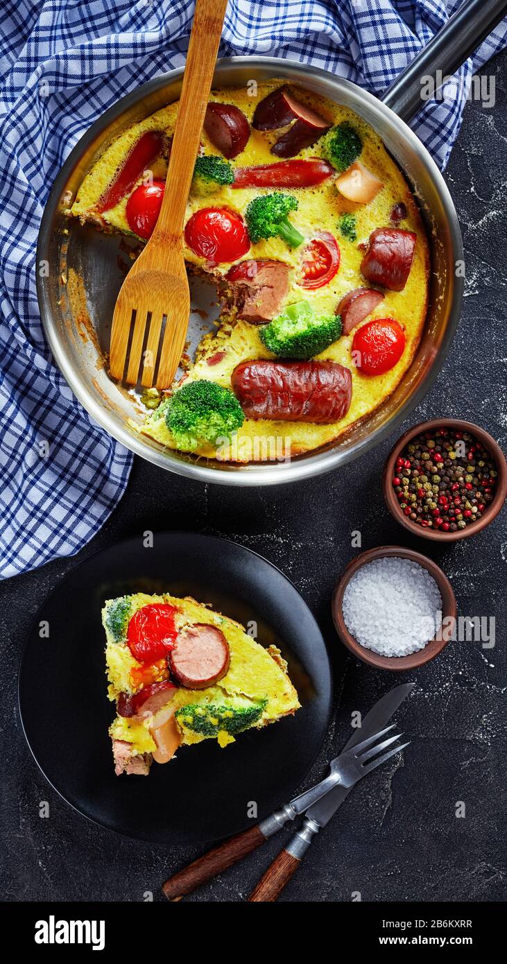 Gebackene Omelette oder Frittata mit Würstchen, Brokkoli, Kirschtomaten, geschmolzenem Käse in einem Skillet auf einem Betontisch und einer Portion auf einem Teller, senkrecht Stockfoto