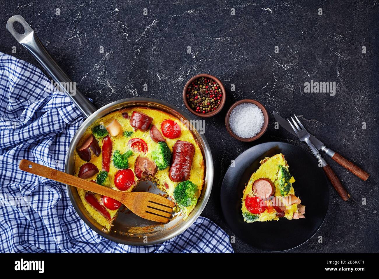 Frittata mit Würstchen, Brokkoli, Kirschtomaten, geschmolzenem Käse in einem Skillet auf einem Betontisch und einem Teil auf einem Teller, horizontale Ansicht von oben Stockfoto